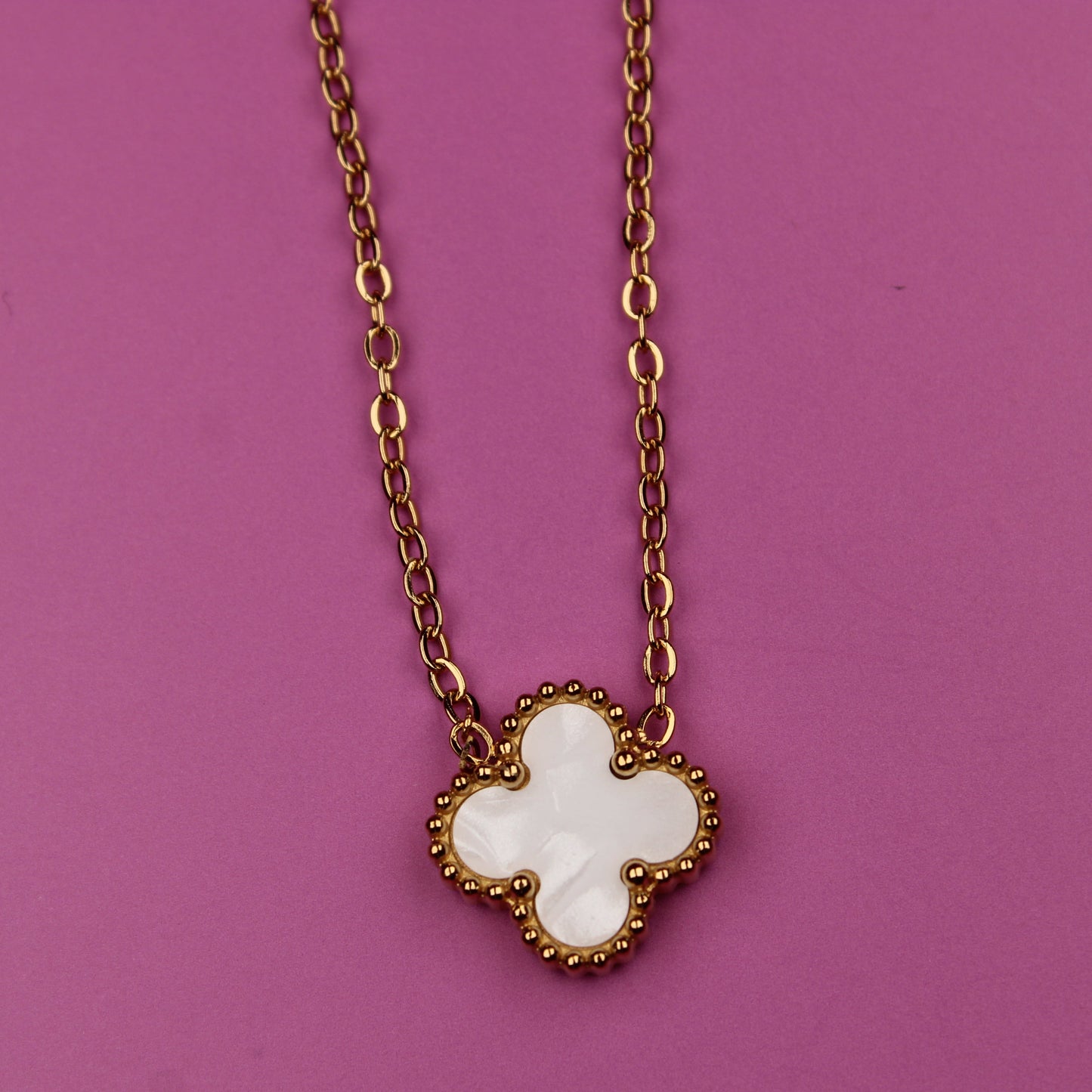 Gold necklace with a white clover-shaped pendant on a pink background