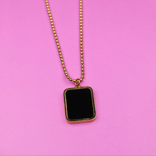 Gold necklace with a black square pendant on a pink background