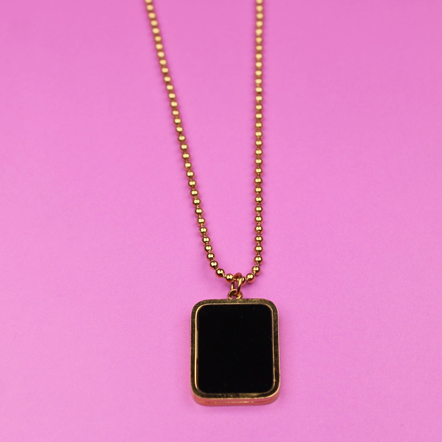 Gold necklace with a black square pendant on a pink background