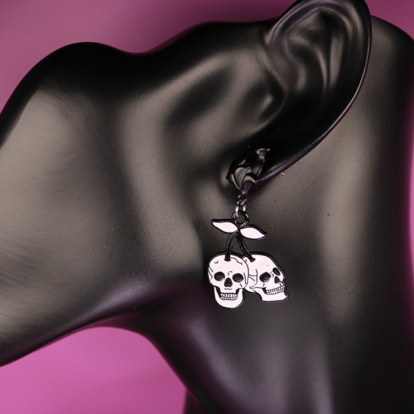 Skull-themed earrings on a black mannequin ear against a pink background