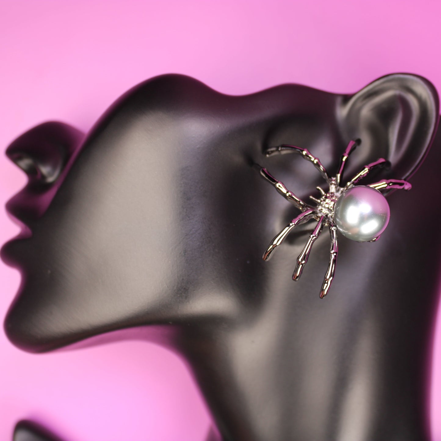 Spider-shaped earring with pearl center on a black mannequin ear against a pink background