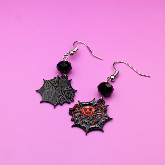 Black spider web earrings with red centers on a pink background