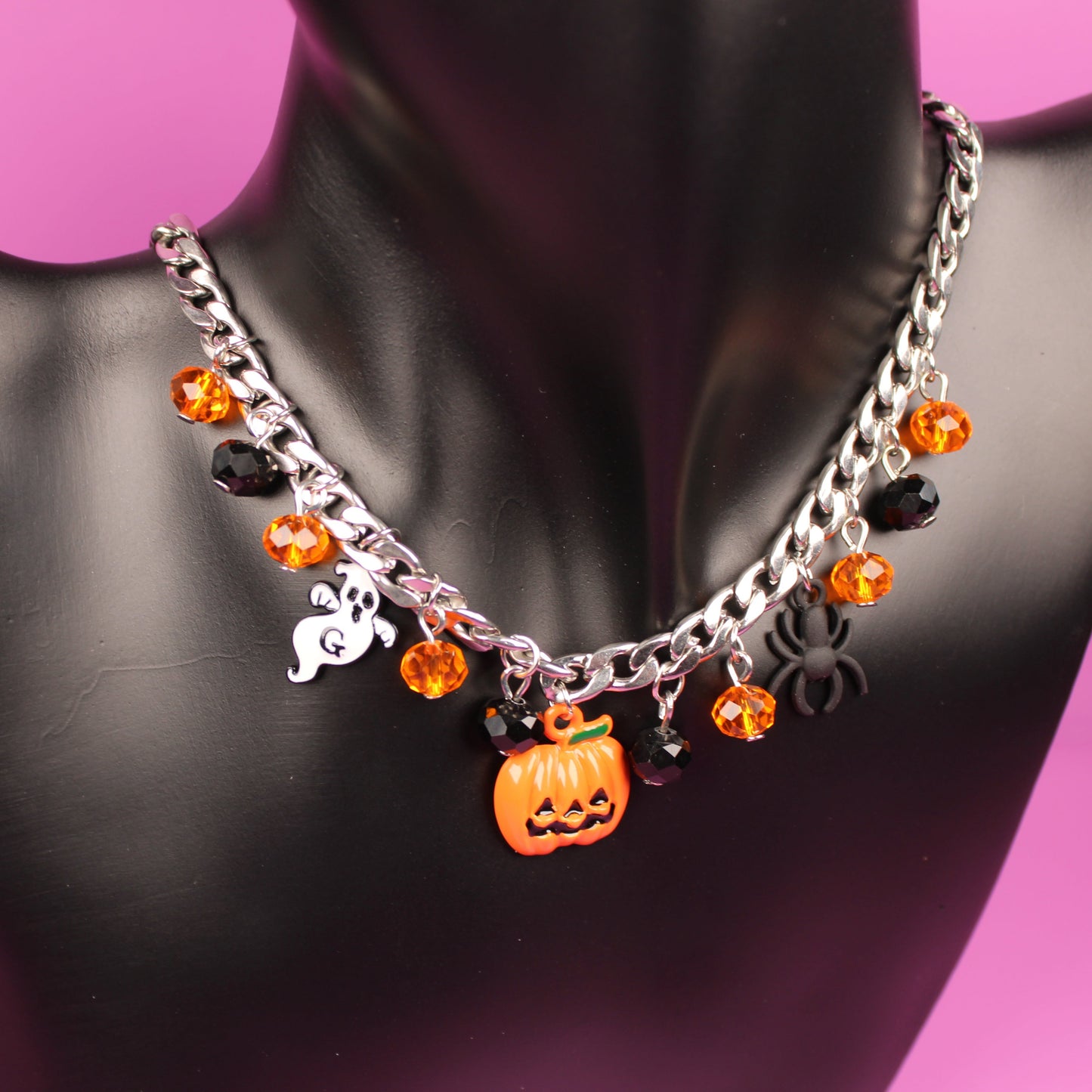 Halloween-themed necklace with pumpkin and ghost charms on a black mannequin against a pink background