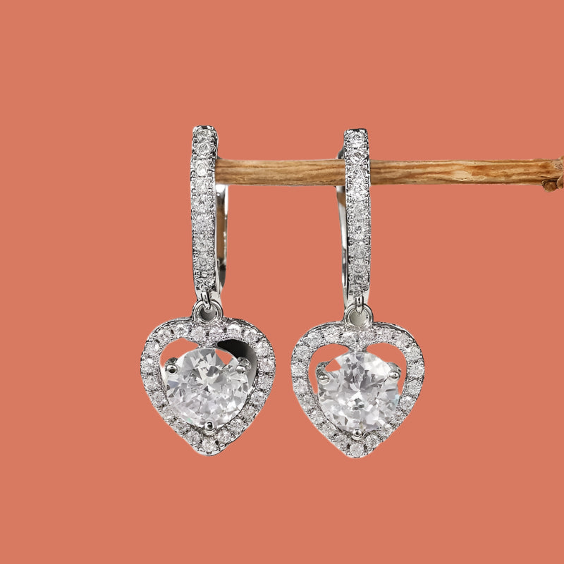 A pair of heart-shaped zircon stud earrings with a silver finish, displayed against a wooden backdrop.
