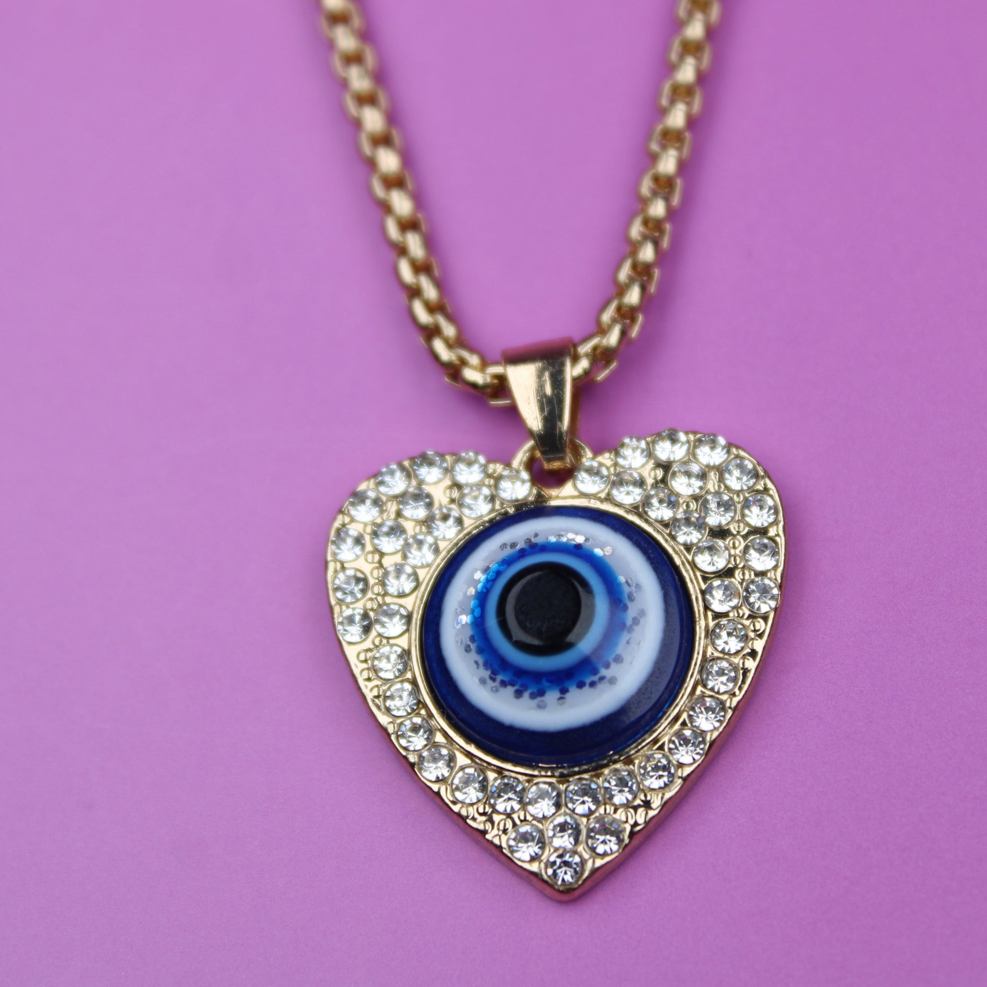 Heart-shaped pendant with a blue eye design on a gold chain against a pink background