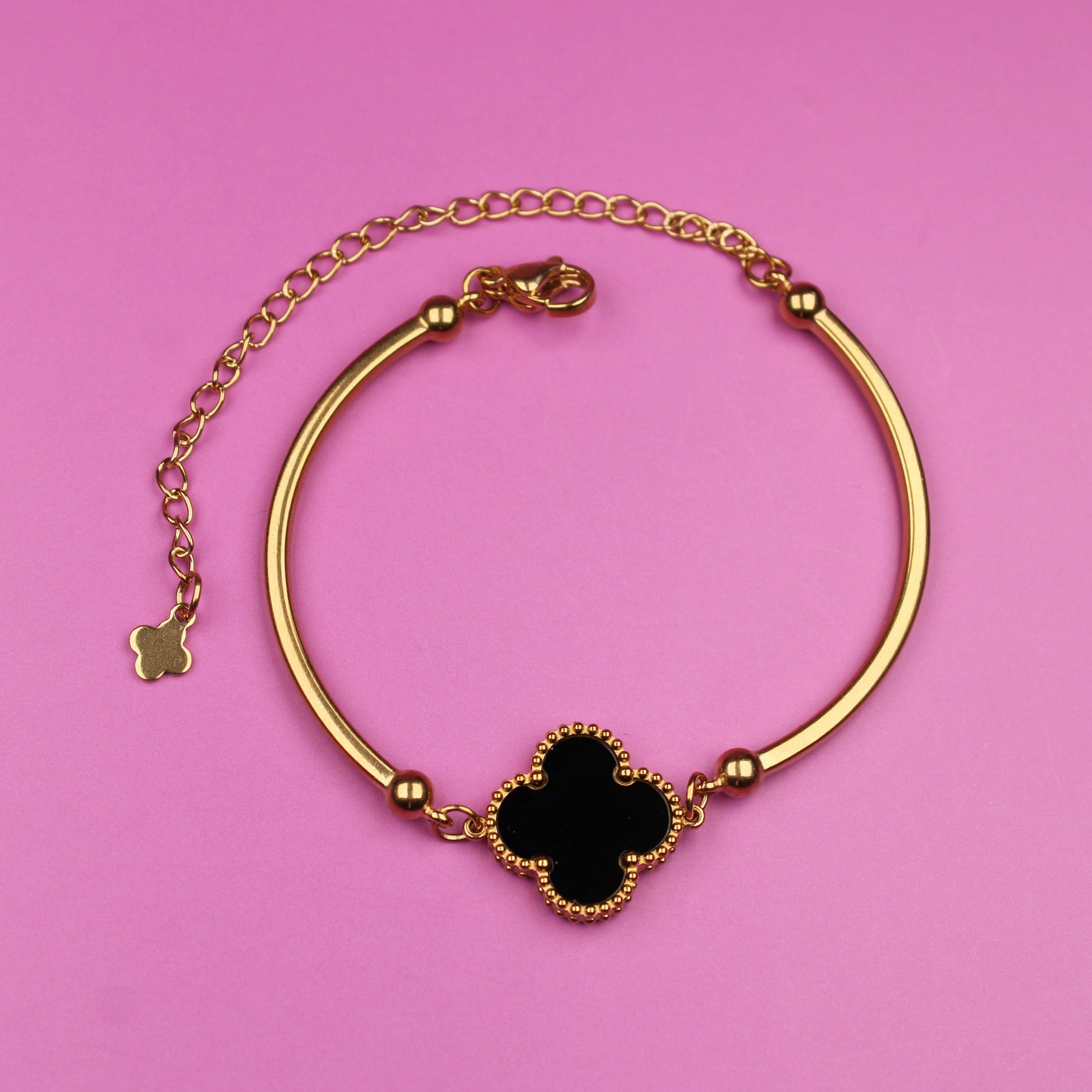 Gold bracelet with a black clover-shaped charm on a pink background