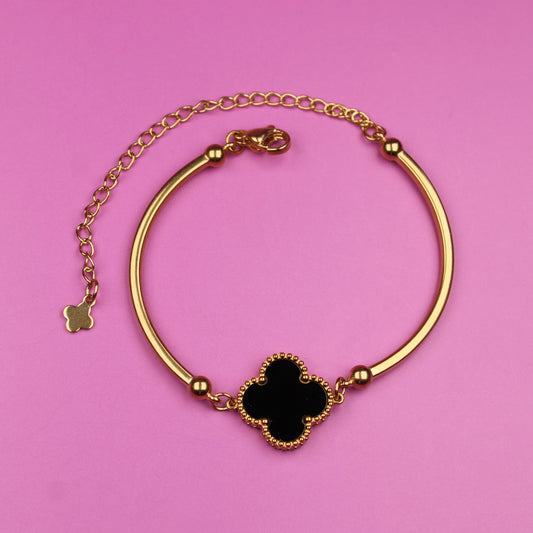 Gold bracelet with a black clover-shaped charm on a pink background