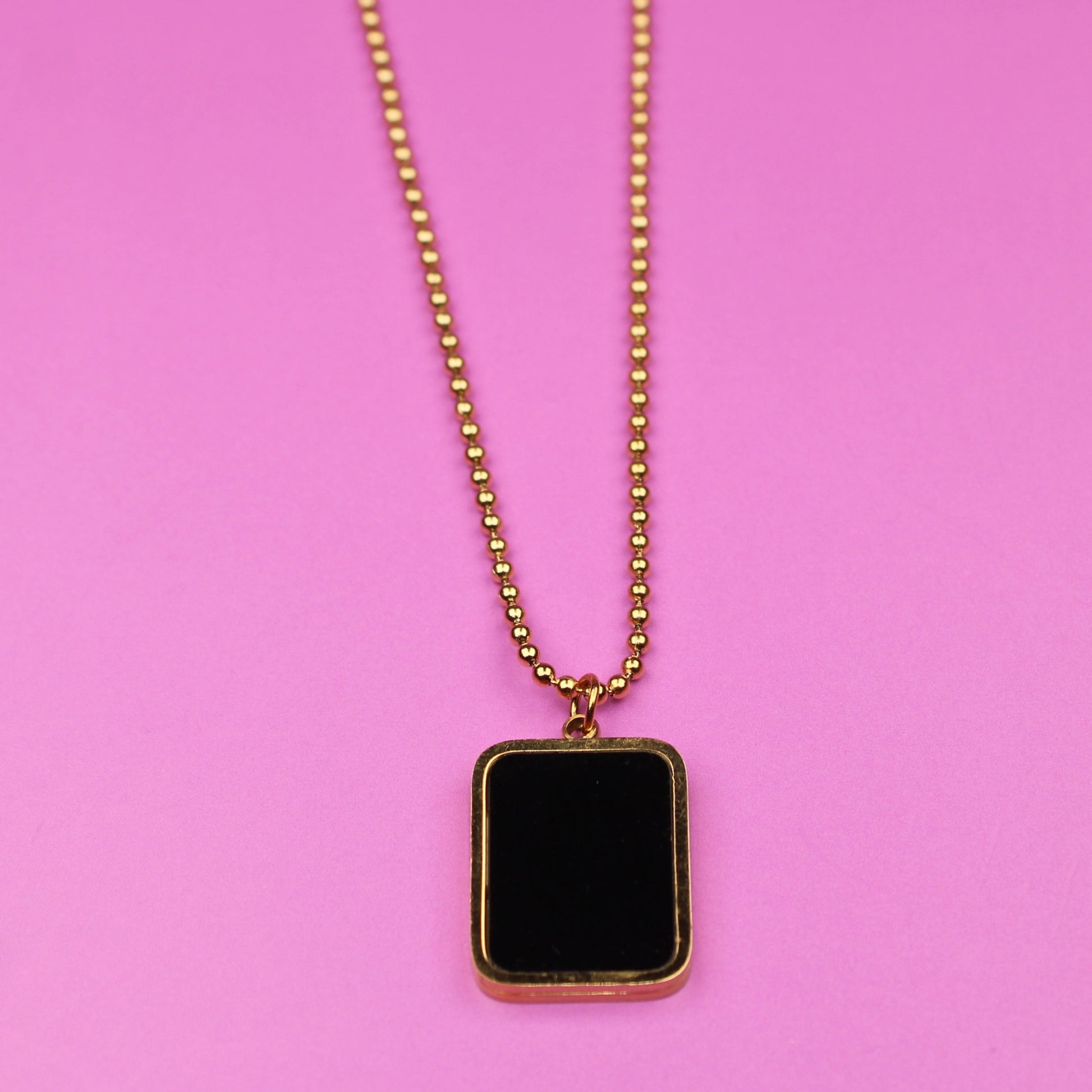 Gold necklace with a black square pendant on a pink background