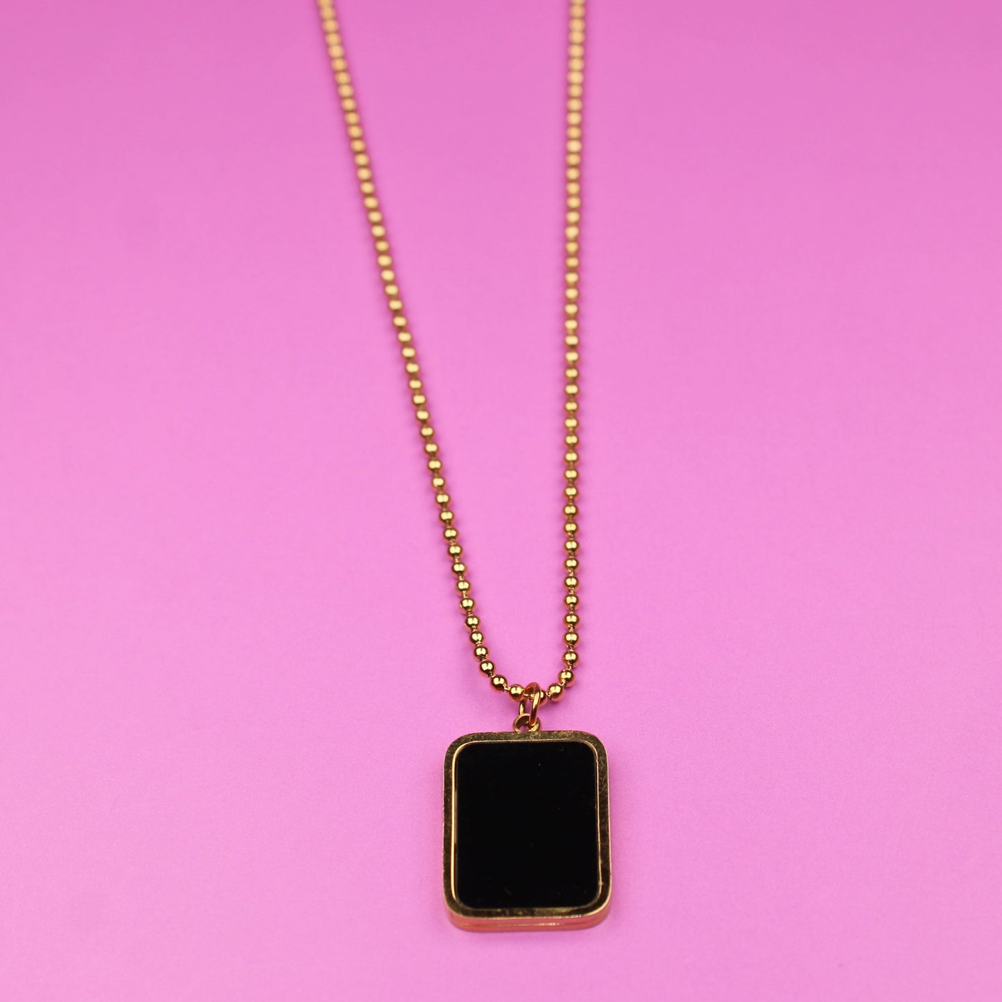 Gold necklace with a black square pendant on a pink background