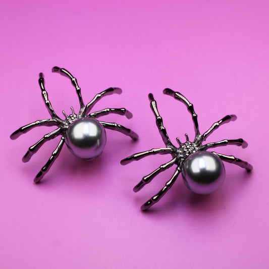 Two spider-shaped earrings with pearl centers on a pink background